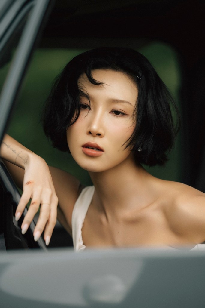 HWASA with short black hair and an elegant pose leaning out of a car window, looking contemplative with a soft expression.