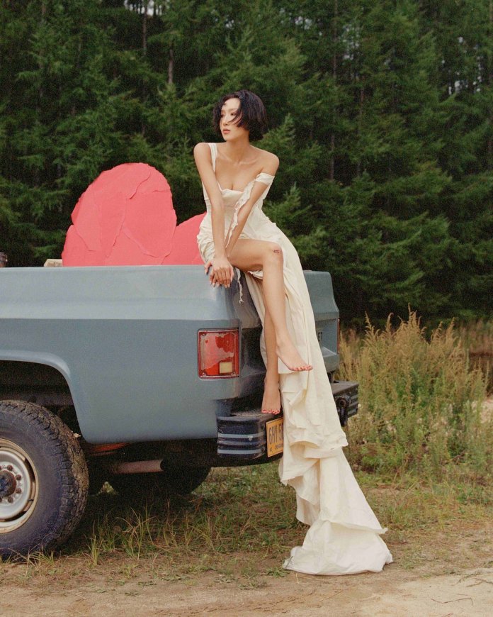 Hwasa flowing white dress poses on the back of a blue pickup truck, with a backdrop of lush green trees and a large pink rock sculpture.