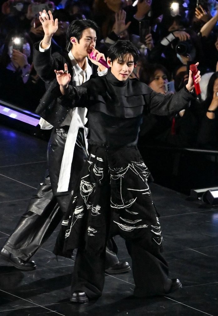 BTS on stage, one holding a microphone and waving, while the other dances enthusiastically. They are dressed in stylish outfits, with a crowd of fans visible in the background.