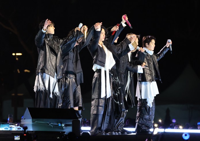 BTS dressed in black outfits on stage, posing with their arms raised, during a concert at night.