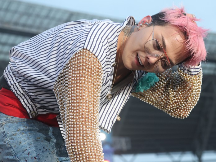 G-Dragon with pink hair wearing a striped shirt with pearl embellishments leans forward while performing on stage.
