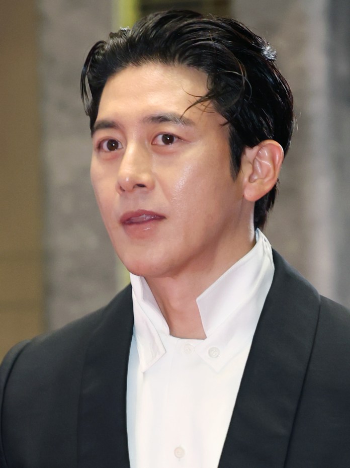 Close-up portrait of Go Soo with dark slicked-back hair, wearing a black suit with a white shirt, looking attentive and slightly smiling.