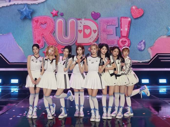 A group of eight female performers in matching white dresses and sporty sneakers, posing together on stage with a colorful backdrop that says 'RUDE!'