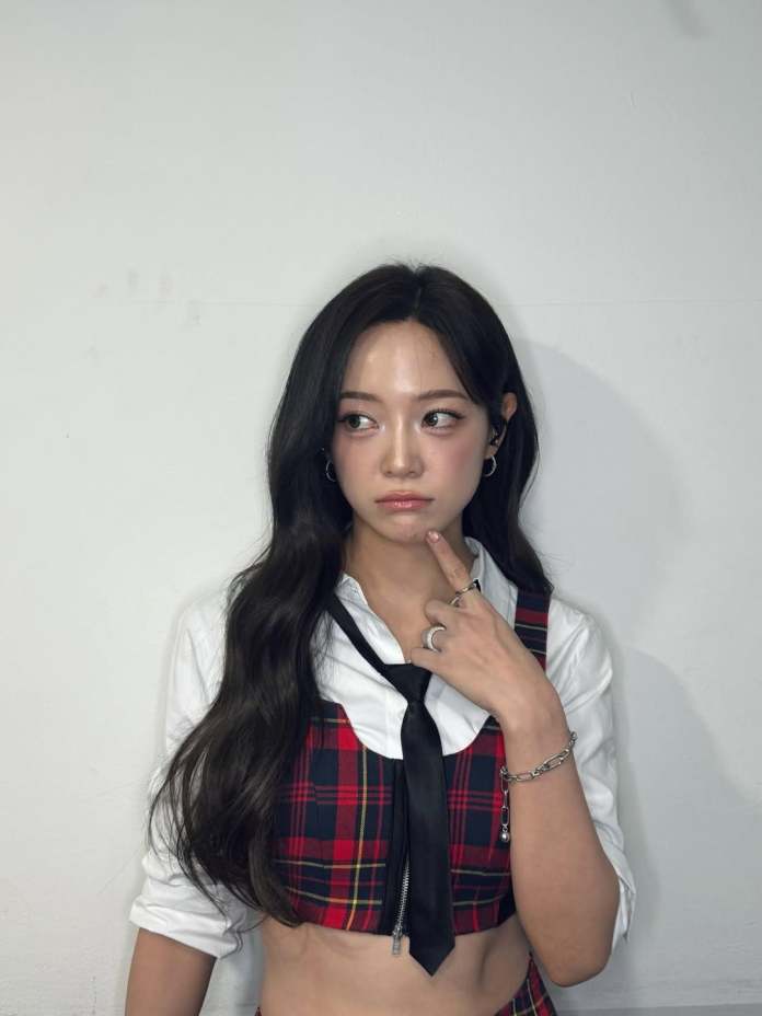 Kim Sejeong with long dark hair, wearing a red plaid crop top with a white shirt underneath and a black tie, poses with a thoughtful expression against a plain white background.