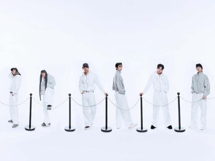 Monsta x dressed in coordinated gray and white outfits stand in a line beside a stanchion and rope barrier, against a plain white background.