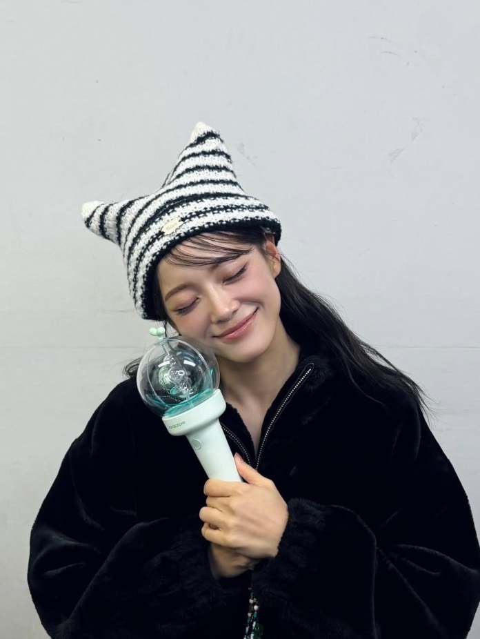 Kim Sejeong  smiling wearing a striped cat-ear hat and a black jacket, holding a lightstick close to their face against a simple gray background.