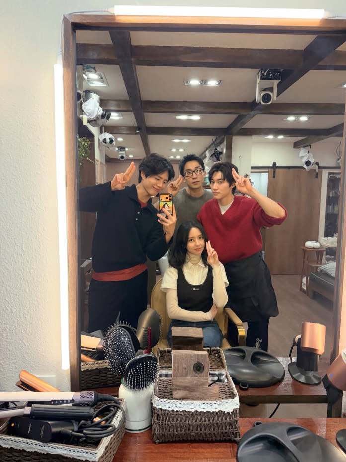 A group of four people posing together in a hair salon, smiling and making peace signs. The scene is reflected in a large mirror, surrounded by various hair styling tools and equipment.