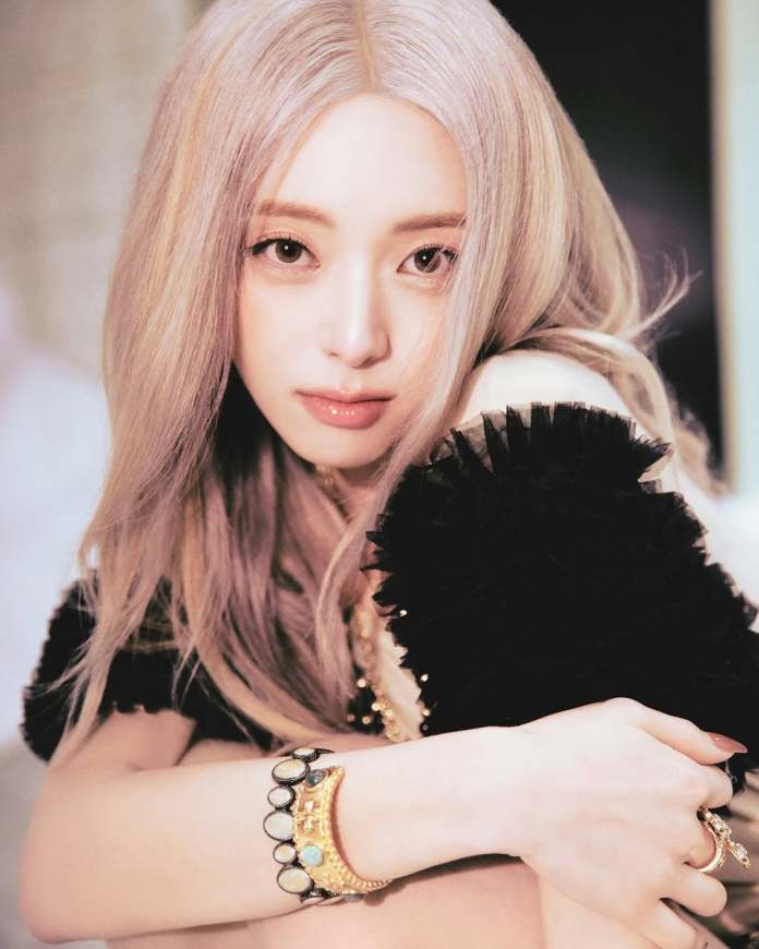 A close-up portrait of Yuna with long, light-colored hair, wearing a black, fluffy top, showcasing a gold bracelet on one wrist.