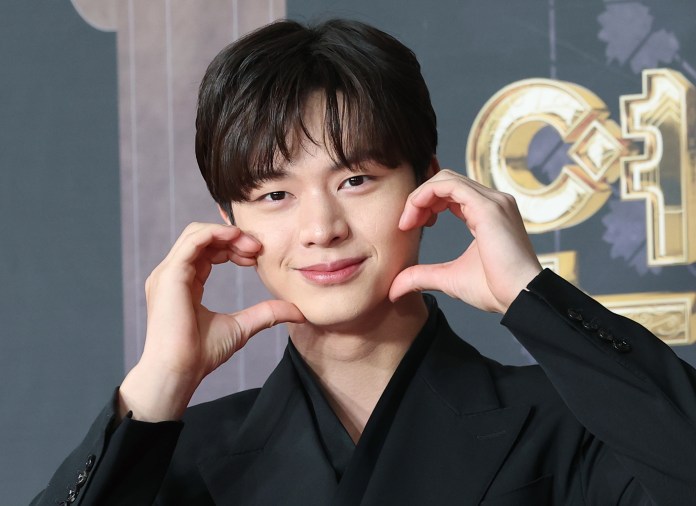 Yook Sungjae in a black suit making a heart shape with his hands and smiling.