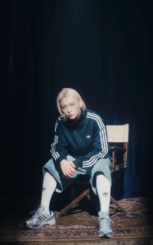Felix sitting on a chair wearing an Adidas tracksuit, knee-length denim shorts, and sneakers, with a contemplative expression, set against a dark background.