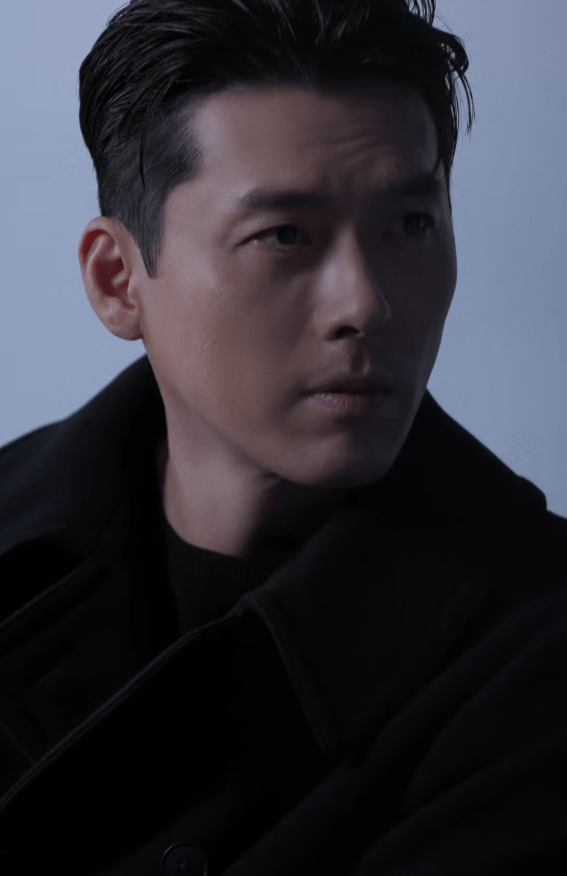 Close-up portrait of Hyun Bin with short, dark hair and a serious expression, wearing a black coat against a soft grey background.