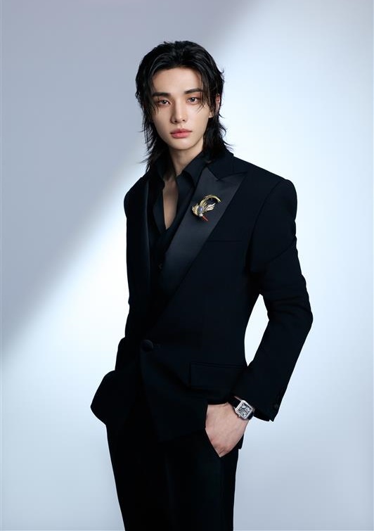 Hyunjin in a formal black suit with a sleek hairstyle, posing confidently against a light gradient background. He wears a black shirt and a decorative brooch on his suit jacket, with his hand resting in his pocket.