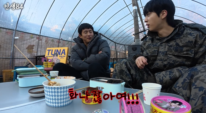 BTS Jin sitting in a greenhouse, engaged in conversation. They are surrounded by various food containers on a table, with one man wearing a camouflage outfit and the other in a puffy jacket. A sign for 'TUNA' is visible in the background.