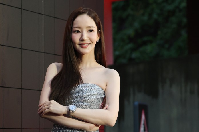 Park Min-young smiling with long hair wearing a sparkling silver dress, standing with her arms crossed in front of a modern outdoor background.