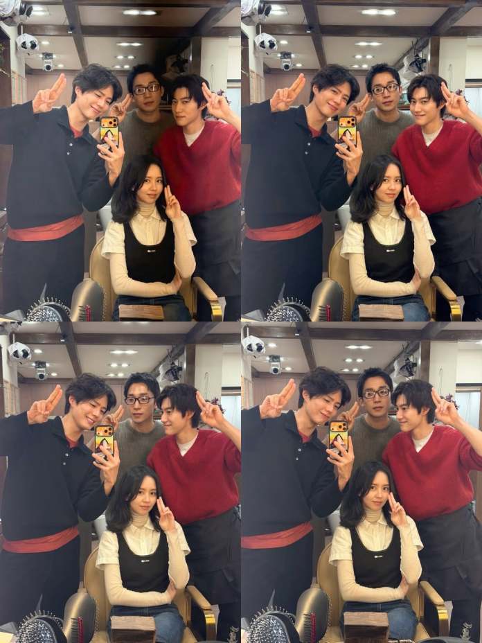 A group of four people posing for a selfie in a makeup room, smiling and making peace signs. Two men are wearing sweaters and a third man is wearing glasses, while a woman in a black dress over a white top sits in front.
