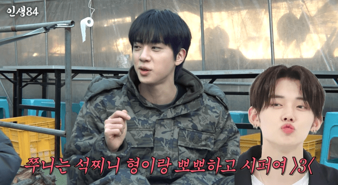 BTS Jin engaged in conversation,  wearing a camouflage jacket and gesturing, while Yeonjun makes a playful pout. Background shows a structure with a light source reflecting off it.