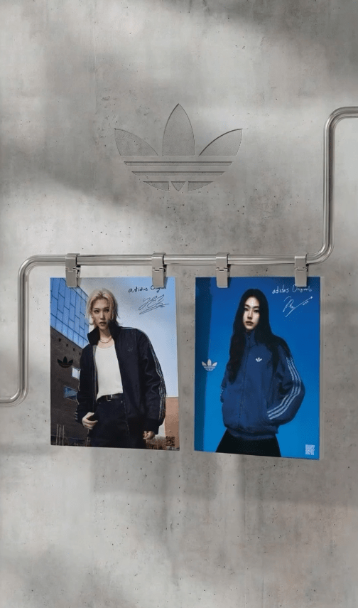 Two promotional posters featuring Felix and Hoyeon in Adidas clothing, hanging on a metal rod against a concrete background. The left poster shows Felix with blonde hair in a black jacket, while the right poster features Hoyeon with dark hair in a blue sweatshirt. Both posters include the Adidas logo.