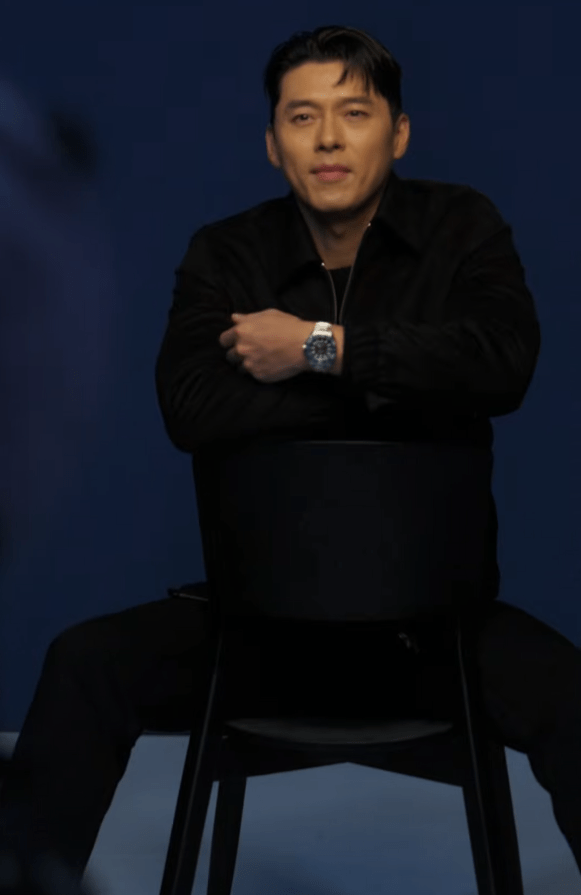 Hyun Bin sitting on a black chair with a blue background, looking directly at the camera and crossing his arms, wearing a black shirt and a watch.