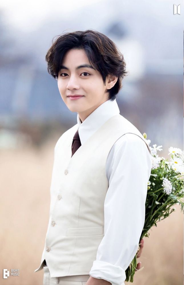 BTS V in a formal outfit, smiling gently while holding a bouquet of white flowers in an outdoor setting.