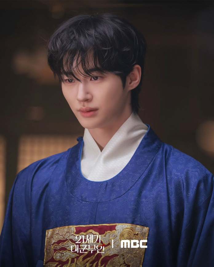 Byeon Woo-seok in a traditional blue hanbok, with a focused expression. He has dark, tousled hair and is set against a softly lit background, suggesting a historical or cultural context.