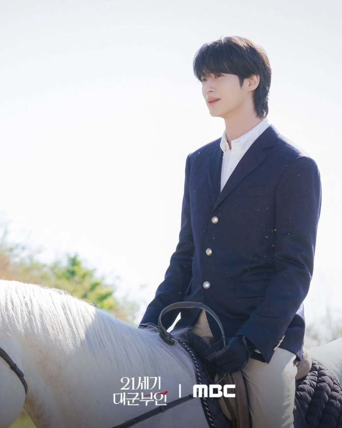Byeon Woo-seok in a navy blazer and white shirt sits on a white horse, gazing thoughtfully into the distance against a bright sky.
