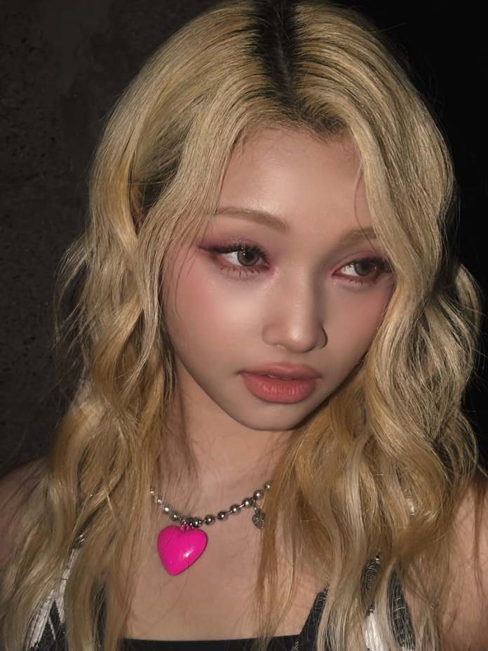 Close-up portrait of Gehlee with long, wavy blonde hair and soft makeup, featuring pink eyeshadow and a heart-shaped pink necklace.