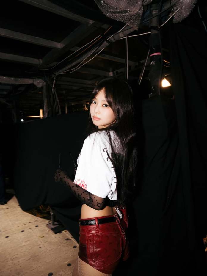 Jennie with long dark hair wearing a white shirt and red shorts, posing in a backstage setting.