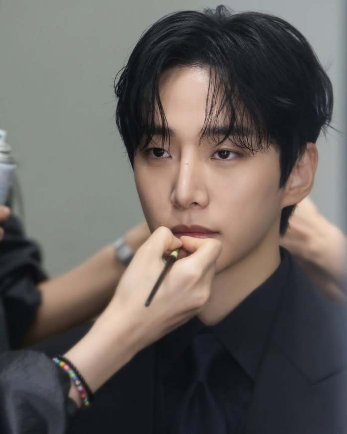 A close-up of Lee Junho with dark hair receiving makeup application, with a makeup artist's hand working on his lips, dressed in a black suit.