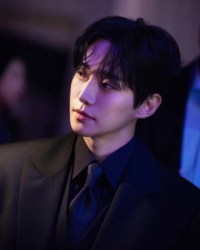 Lee Junho in a black suit and tie with tousled dark hair, gazing thoughtfully to the side.