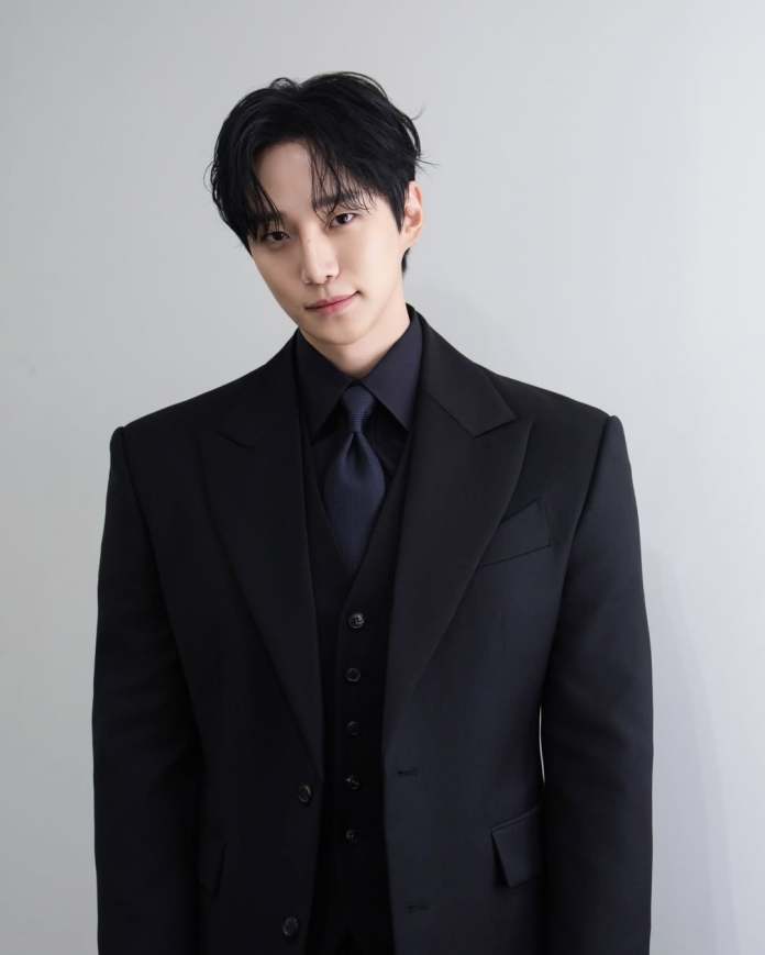 Lee Junho wearing a formal black suit and tie, standing against a neutral background, looking directly at the camera with a slight smile.