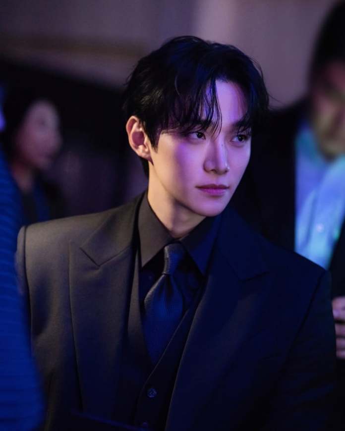 Lee Junho with dark hair and formal attire looks thoughtfully away from the camera in a dimly lit setting.