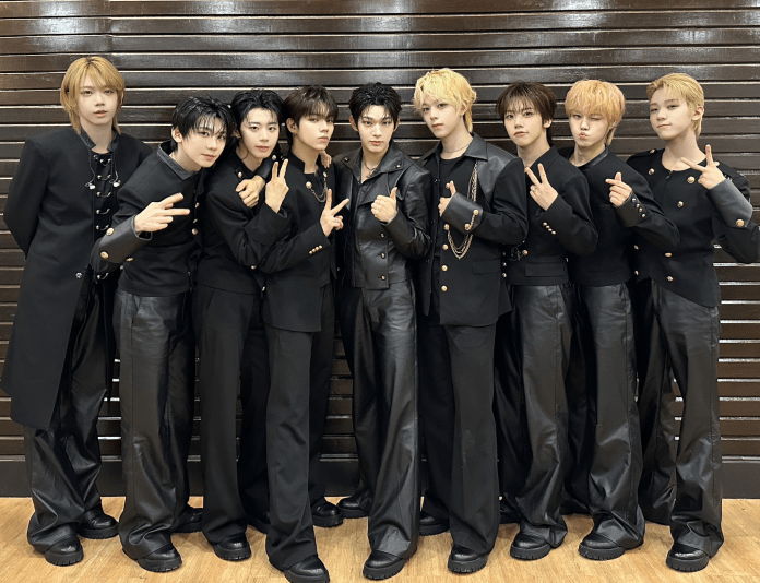 AHOF posing together in stylish matching black outfits with unique designs, standing in front of a wooden backdrop.
