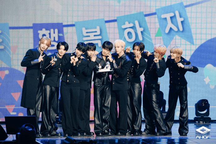 AHOF poses together on stage, dressed in matching black outfits. They are holding a small cake, smiling, and making playful hand gestures against a colorful backdrop with cheerful decorations and text.