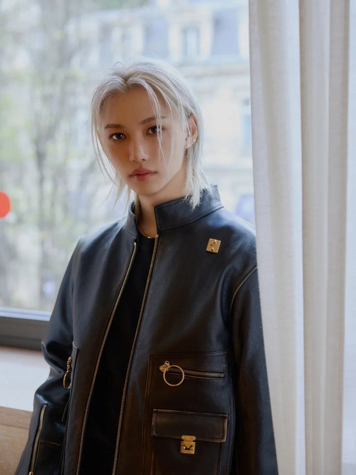 Felix with shoulder-length platinum blonde hair wearing a stylish black leather jacket with decorative zippers and pockets, looking directly at the camera with a neutral expression, standing near a window with soft natural light.