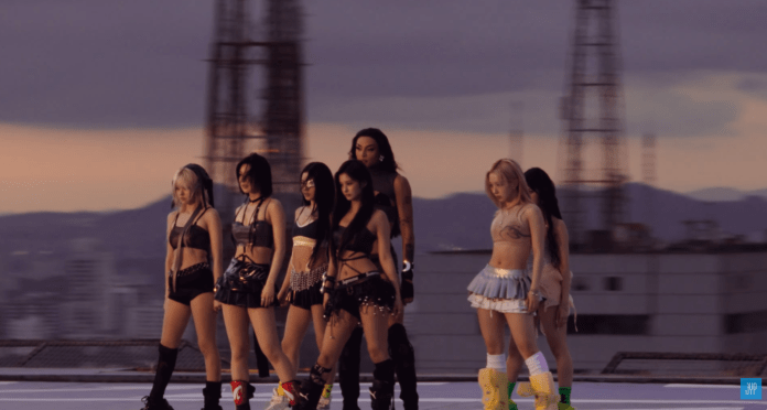 NMIXX and Pabllo Vittar in stylish outfits standing together on a rooftop during sunset, with cityscape and antennas in the background.