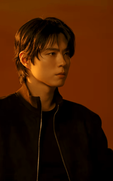 A close-up portrait Park Bo-gum with dark, slightly wavy hair, wearing a black jacket, set against an orange background. He has a serious expression and is looking to the side.