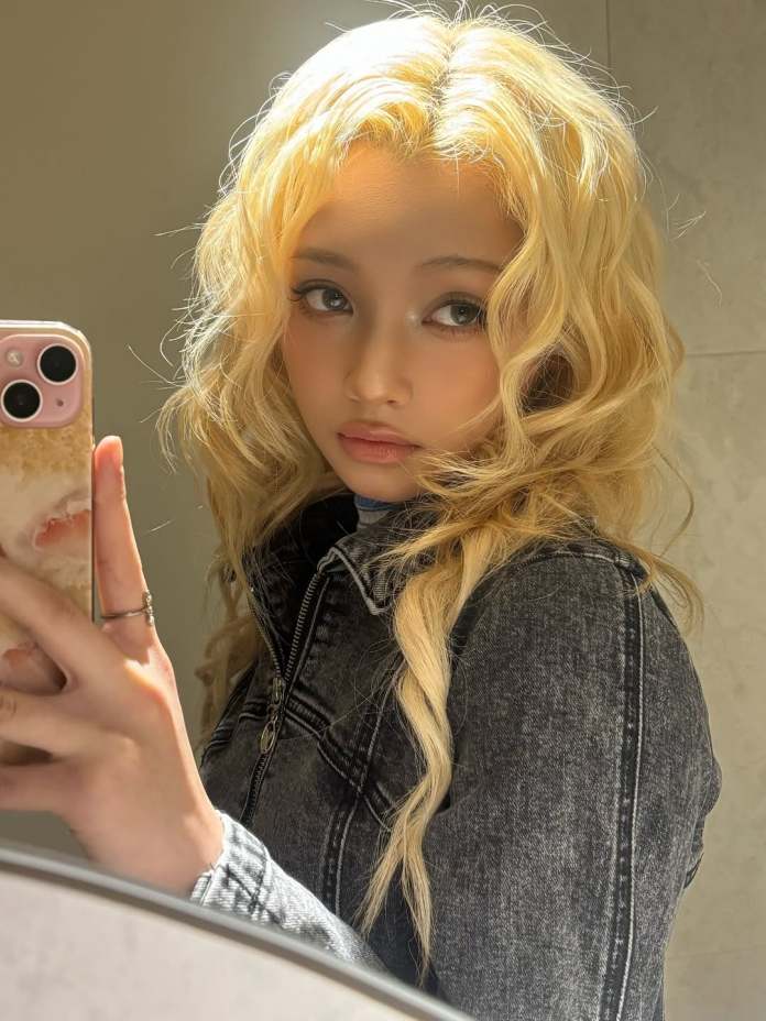 Gehlee with long, wavy blonde hair, wearing a denim jacket, poses for a selfie in front of a mirror, holding a smartphone.