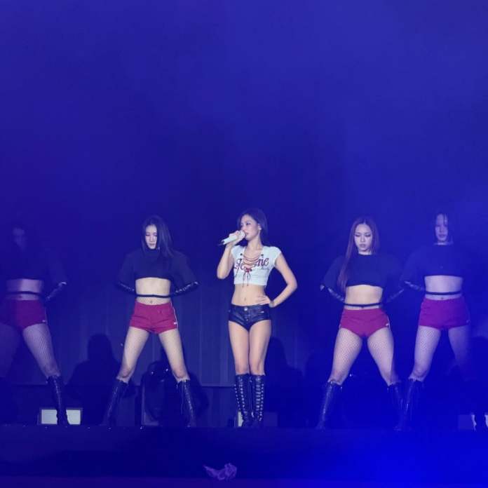 A female singer performs on stage, dressed in a cropped top and shorts, surrounded by four backup dancers in black tops and red shorts.