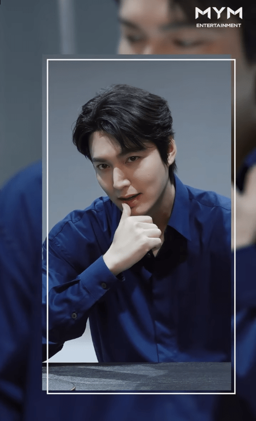 Lee Minho with dark hair poses thoughtfully, resting their chin on their hand while wearing a blue shirt. The background is softly blurred, focusing attention on the individual.
