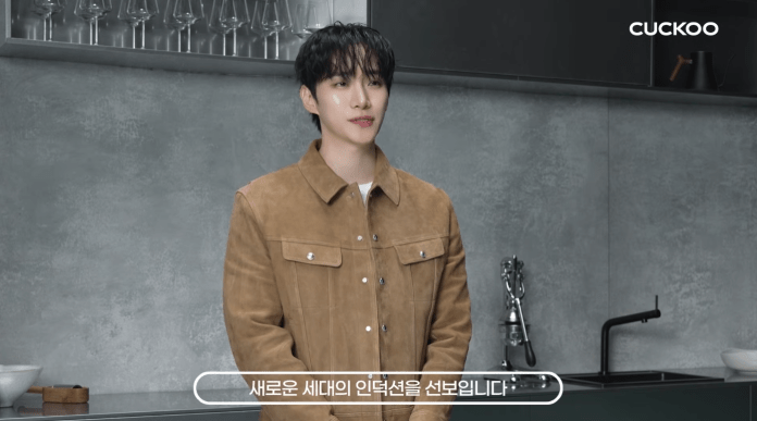 Lee Junho standing in a modern kitchen, wearing a brown suede shirt and looking at the camera, with various kitchen utensils and glasses in the background.