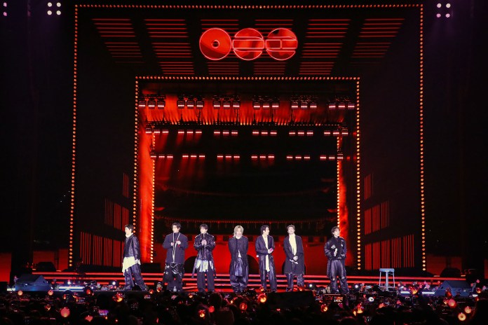 BTS performing on stage with dramatic red lighting and a large backdrop, surrounded by fans holding light sticks.