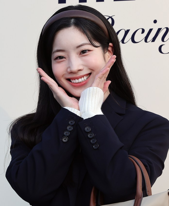 A woman in a dark blazer and white sweater sleeves smiles and poses playfully with her hands on her cheeks, showcasing a happy expression.