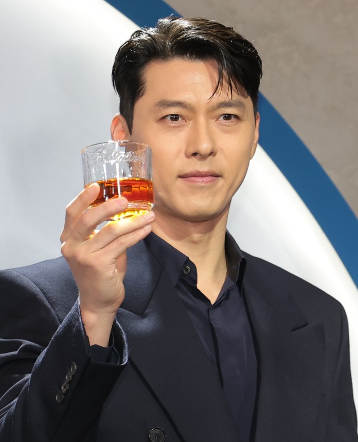 Hyun bin holding up a glass of whiskey, dressed in a dark blazer, against a neutral background.