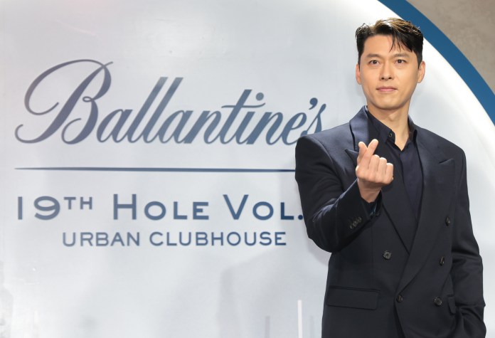 Hyun Bin in a dark suit is smiling and making a hand gesture in front of a branded backdrop that reads 'Ballantine's 19th Hole Vol. Urban Clubhouse.'