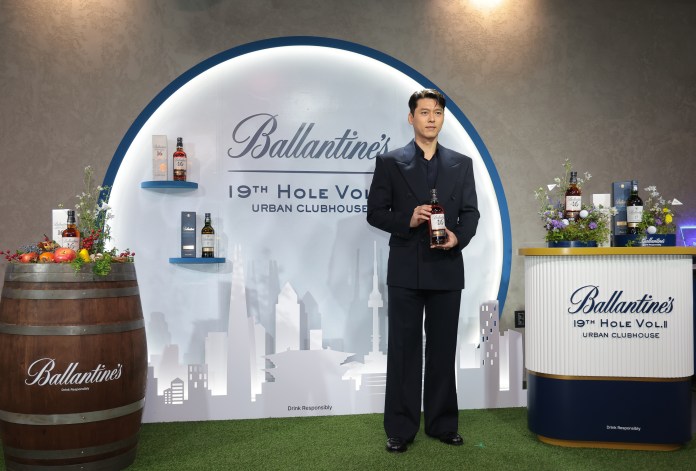 Hyun bin in a stylish black suit holds a bottle of Ballantine's whiskey in front of a backdrop featuring the brand's logo and selection of whiskey bottles on display.