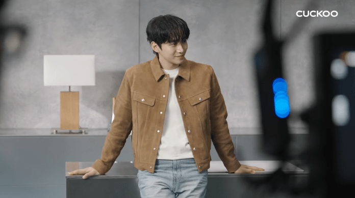 Lee Junho in a brown suede jacket and white shirt stands casually in a modern interior with a lamp and a counter, smiling slightly.