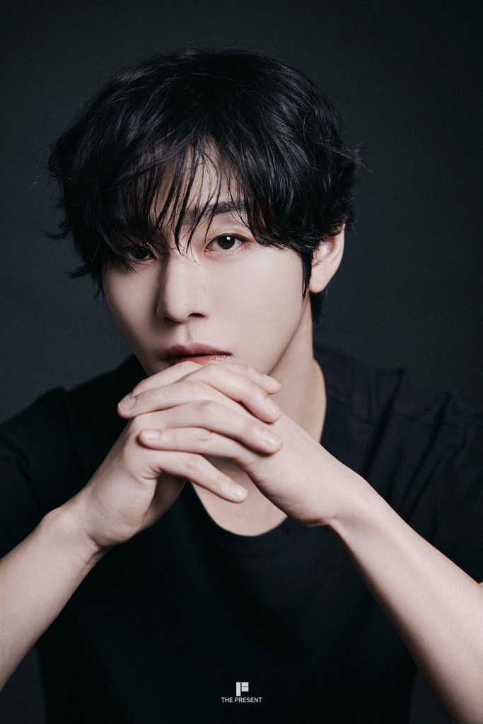 A close-up portrait of Ahn Hyo-seop with dark, tousled hair, wearing a black t-shirt. He is gazing directly at the camera with his hands clasped together in front of his face.