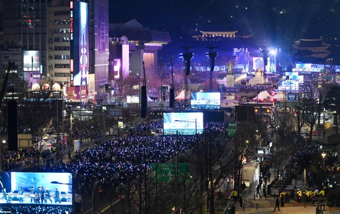A large crowd for BTS gathers at night for an event, illuminated by colorful lights and multiple large screens displaying performances. Skyscrapers and historical buildings are visible in the background.