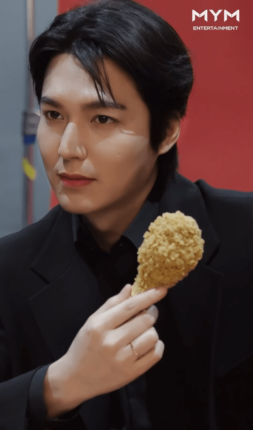Lee Minho in a black suit holding a piece of fried chicken, looking thoughtfully off to the side.