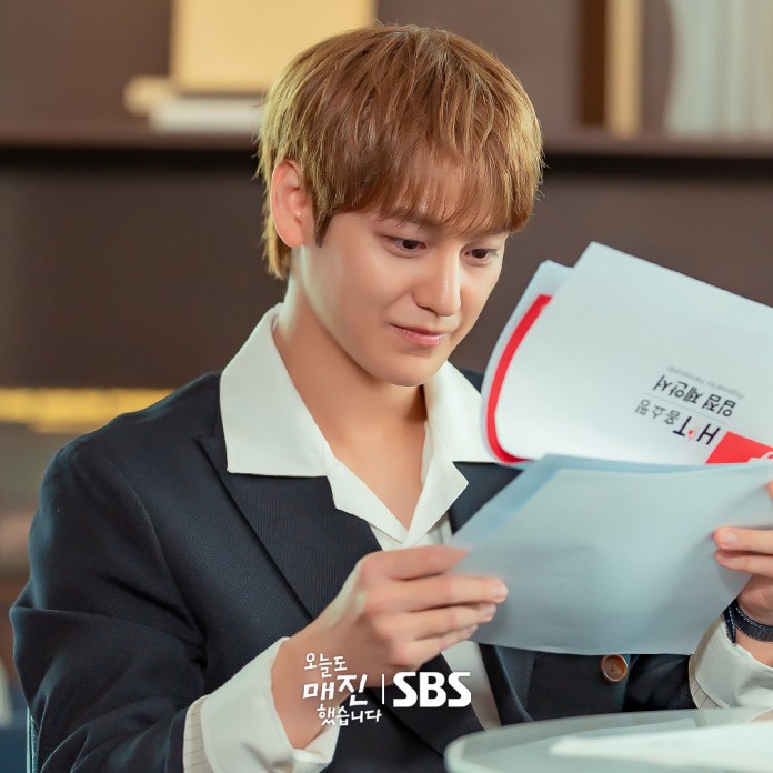 Kim Bum with light brown hair, dressed in a dark suit with a white collar, is sitting at a table and looking thoughtfully at several papers in his hands.